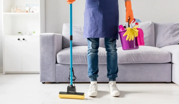 Person in an apron holding cleaning tools, including a mop and a bucket with cleaning supplies, standing in a modern living room, illustrating professional house cleaning services.
